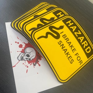 Peut inclure: Plusieurs autocollants d'avertissement jaunes et noirs avec le texte "HAZARD I BRAKE FOR SNAKES". Une carte blanche avec un motif de crâne et des éclaboussures rouges est partiellement visible.