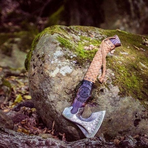 May include: A handcrafted axe with a silver blade featuring intricate carvings. The handle is wrapped in woven tan leather, with a dark purple section and a decorative emblem. The axe rests on a moss-covered rock in a forest setting.