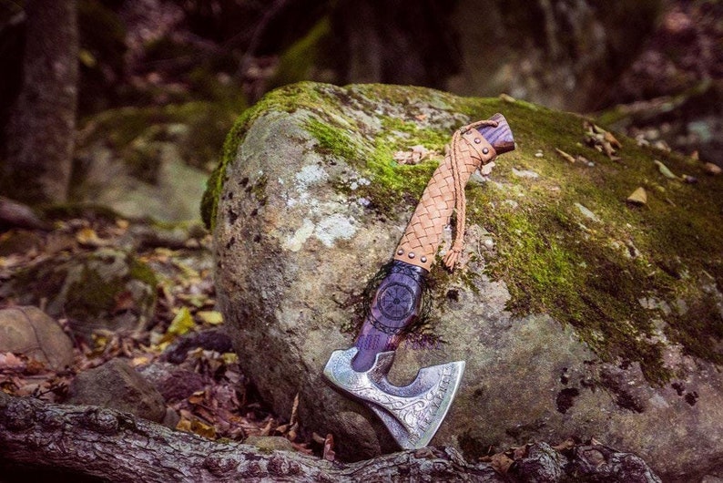 May include: A handcrafted axe with a silver blade featuring intricate carvings. The handle is wrapped in woven tan leather, with a dark purple section and a decorative emblem. The axe rests on a moss-covered rock in a forest setting.