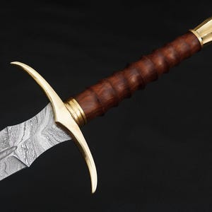 May include: A sword with a wooden handle and a gold-colored hilt. The blade is made of Damascus steel and has a wavy pattern.