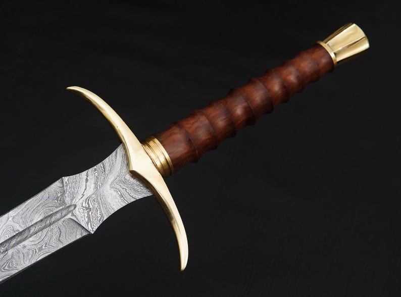 May include: A sword with a wooden handle and a gold-colored hilt. The blade is made of Damascus steel and has a wavy pattern.