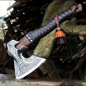 May include: A silver axe with a leather handle and a tassel. The axe head has intricate carvings and a braided leather wrap around the handle.