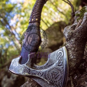 May include: A silver and black axe with intricate carvings, featuring a leather-wrapped handle and a lanyard. The axe head displays a circular emblem and the text "GODS FORCE". The axe is resting on a tree branch.