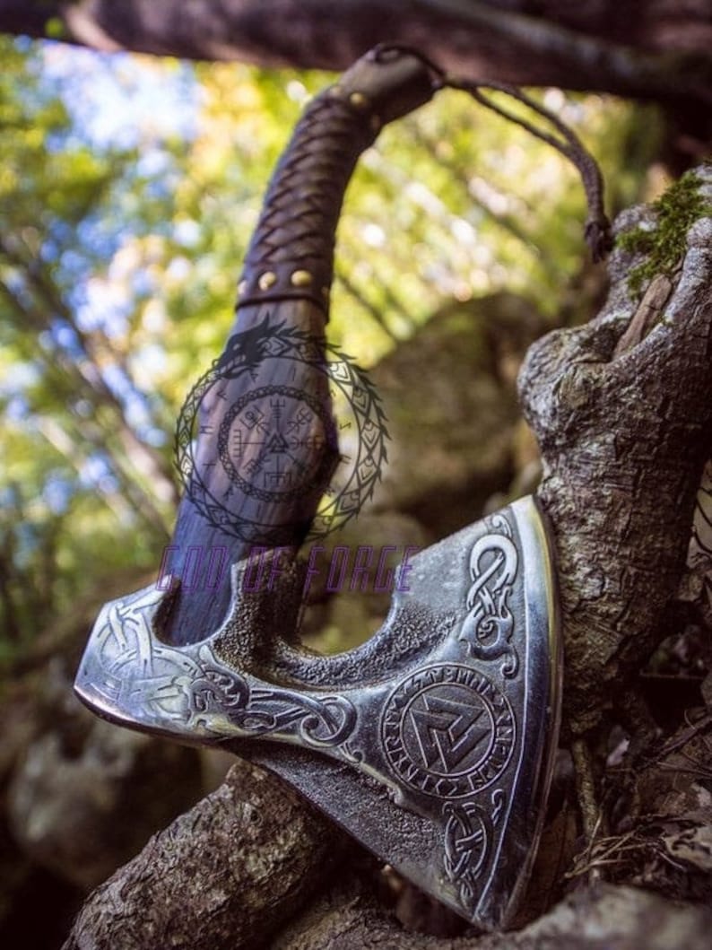 May include: A silver and black axe with intricate carvings, featuring a leather-wrapped handle and a lanyard. The axe head displays a circular emblem and the text "GODS FORCE". The axe is resting on a tree branch.
