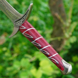 May include: Close-up of a sword hilt with a red and silver wood-grain pattern. The hilt is made of metal and has a decorative design.
