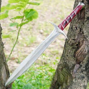 May include: A silver sword with a red and black handle. The sword has a decorative design on the blade and a pointed tip. The sword is resting on a tree trunk.