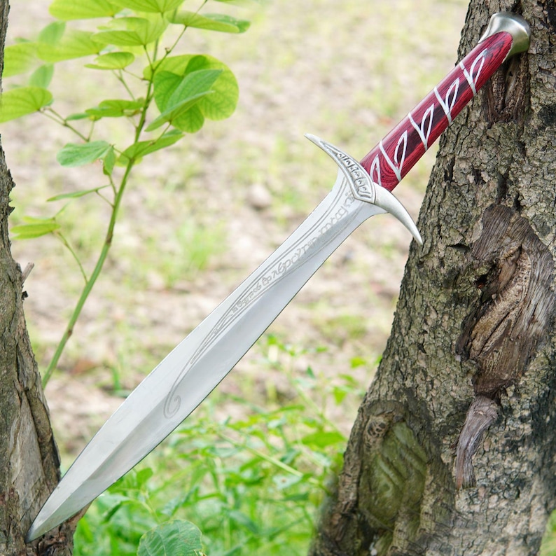 May include: A silver sword with a red and black handle. The sword has a decorative design on the blade and a pointed tip. The sword is resting on a tree trunk.