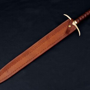 May include: A brown leather scabbard with a decorative pattern and a gold-colored metal tip. The scabbard is for a sword with a brown leather wrapped handle and a gold-colored metal pommel.