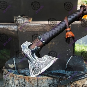 May include: A detailed close-up of a handcrafted axe with a silver blade and a dark wooden handle wrapped in black leather. The axe rests on a black anvil, showcasing its intricate design and craftsmanship. The axe has a brown leather strap and orange tassel.