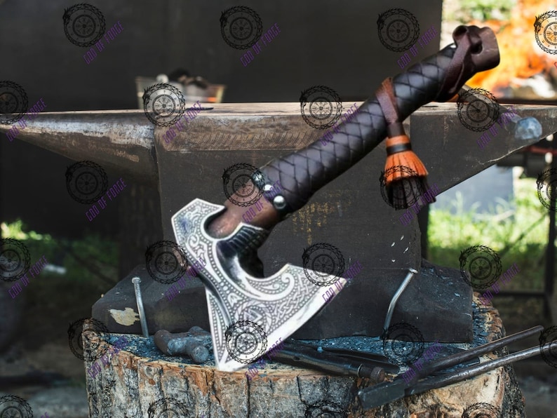 May include: A detailed close-up of a handcrafted axe with a silver blade and a dark wooden handle wrapped in black leather. The axe rests on a black anvil, showcasing its intricate design and craftsmanship. The axe has a brown leather strap and orange tassel.