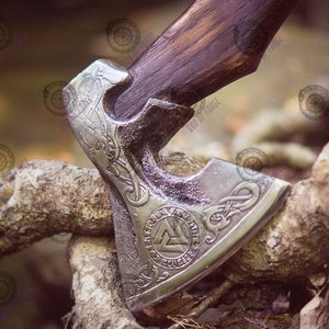 May include: A close-up of a Viking axe with a wooden handle and a silver-colored head. The axe head features intricate engraved designs, including a triquetra symbol. The axe is resting on a weathered wooden surface.