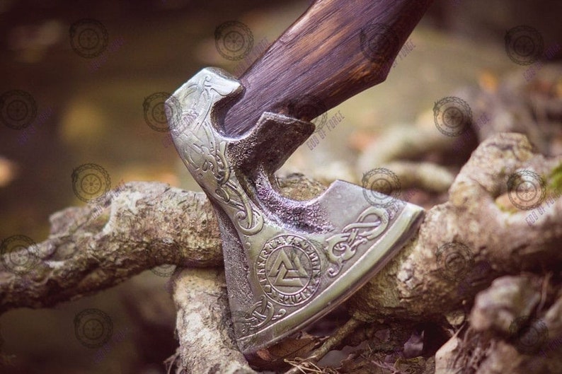 May include: A close-up of a Viking axe with a wooden handle and a silver-colored head. The axe head features intricate engraved designs, including a triquetra symbol. The axe is resting on a weathered wooden surface.