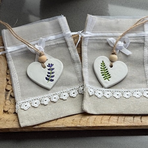 May include: Two small, beige linen sachets with white lace trim and heart-shaped ceramic pendants. One pendant features a lavender sprig, the other a fern. Each sachet has a jute string and wooden bead.