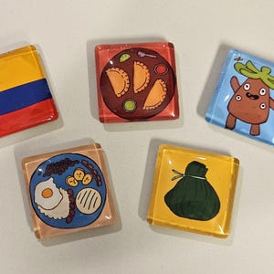 Puede incluir: Cinco imanes cuadrados de vidrio con diseños distintos: uno con la bandera colombiana amarilla, azul y roja, uno con tres empanadas en un plato, uno con un personaje de dibujos animados, uno con un plato de desayuno y uno con un tamal verde.
