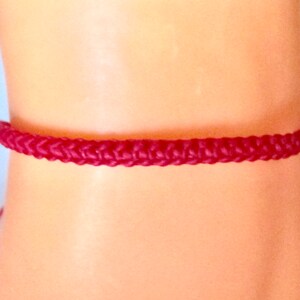 May include: A burgundy braided bracelet is displayed on a wrist. The bracelet is made of a woven material and has a simple, minimalist design. The bracelet is a deep red colour and appears to be adjustable.