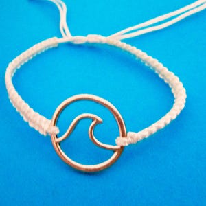 May include: A white macrame bracelet featuring a gold-coloured wave charm. The wave design is an outline within a circular frame. The bracelet is set against a bright blue background.