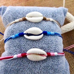 May include: Three seashell bracelets with adjustable cord fastenings. The bracelets feature white seashells and cords in shades of brown, blue, and pink. They are displayed on a light blue surface with a starfish in the background.