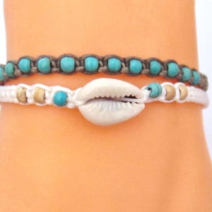 May include: Two handmade bracelets. The top bracelet is brown with turquoise beads. The bottom bracelet is white with a seashell and turquoise and tan beads. The bracelets are on a light skin tone.