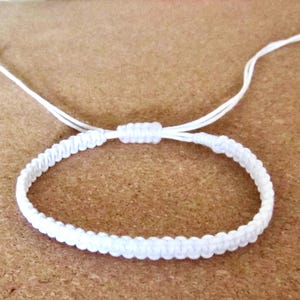 May include: A white macramé bracelet with an adjustable sliding knot closure. The bracelet is made of woven white cord and is displayed on a light brown surface. The bracelet's design is simple and elegant, suitable for everyday wear.