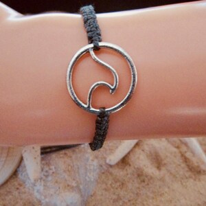May include: A silver-coloured bracelet with a wave design inside a circle, attached to a dark grey woven cord. The bracelet is shown on an arm, with a sandy background.