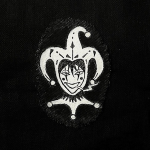 May include: A black patch with a white jester design. The jester has a smiling face, a tall hat with bells, and is smoking a cigarette. The patch is oval-shaped and has a stitched border.