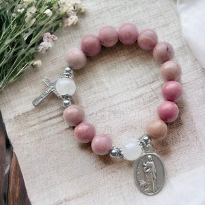 May include: A pink and white beaded bracelet with a silver cross charm and a silver medal charm featuring a religious figure. The bracelet is made of natural stones and is perfect for a spiritual or religious gift.