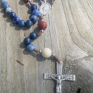 May include: A rosary with blue, red, and cream-colored beads. The beads are strung on a brown cord and separated by silver-colored metal beads. A silver crucifix and a medal with a saint's image are at the end.