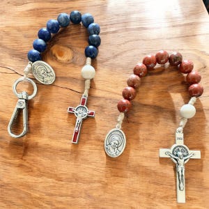 May include: Two rosaries with wooden beads. One rosary has blue beads and a silver cross with a figure of Jesus Christ. The other rosary has red beads and a silver cross with a figure of Jesus Christ. Both rosaries have a silver charm with a figure of a saint.