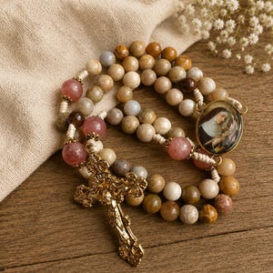 May include: A rosary featuring round beads in shades of tan, brown, and translucent pink, strung on light-colored cord. It includes a gold-toned metal crucifix with ornate detailing and an oval pendant depicting a religious figure in a habit.
