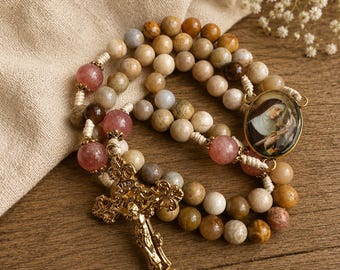 Pode incluir: Um rosário com contas redondas em tons de bege, castanho e rosa translúcido, enfiadas num cordão claro. Inclui um crucifixo de metal dourado com detalhes ornamentados e um pendente oval que representa uma figura religiosa em hábito.