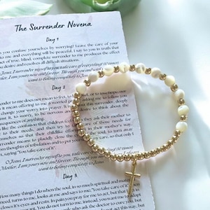 May include: A gold bracelet with white beads and a gold cross charm. The bracelet is on top of a page with the text "The Surrender Novena" and the text "Jesus, I surrender myself to you take care of everything (10 times)".