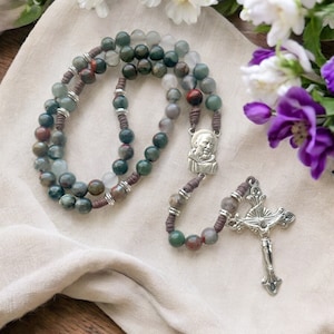 May include: A brown and green stone rosary with a silver crucifix and a silver charm of a man's face. The rosary is on a white fabric background.