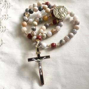 May include: A rosary with multi-colored stone beads, including white, gray, brown, and red. The rosary features a gold-colored metal crucifix with a silver figure of Jesus and a circular medallion with a religious figure.