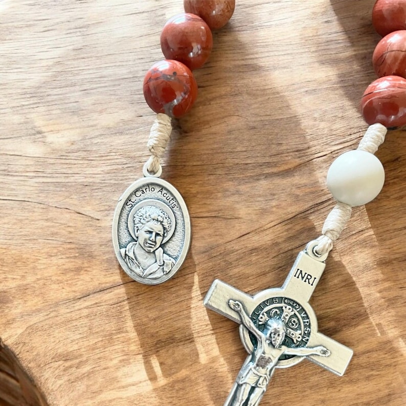 May include: A brown and white rosary with a silver crucifix and a silver medal featuring a picture of St. Carlo Acutis. The medal has the text "St. Carlo Acutis" around the edge. The crucifix has the text "INRI" on the top.
