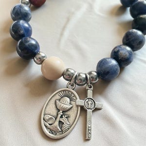 May include: A beaded bracelet featuring dark blue and cream-colored beads. The bracelet includes a silver-toned cross charm and an oval charm with a chalice and grapes. The word "Italy" is inscribed on the oval charm.