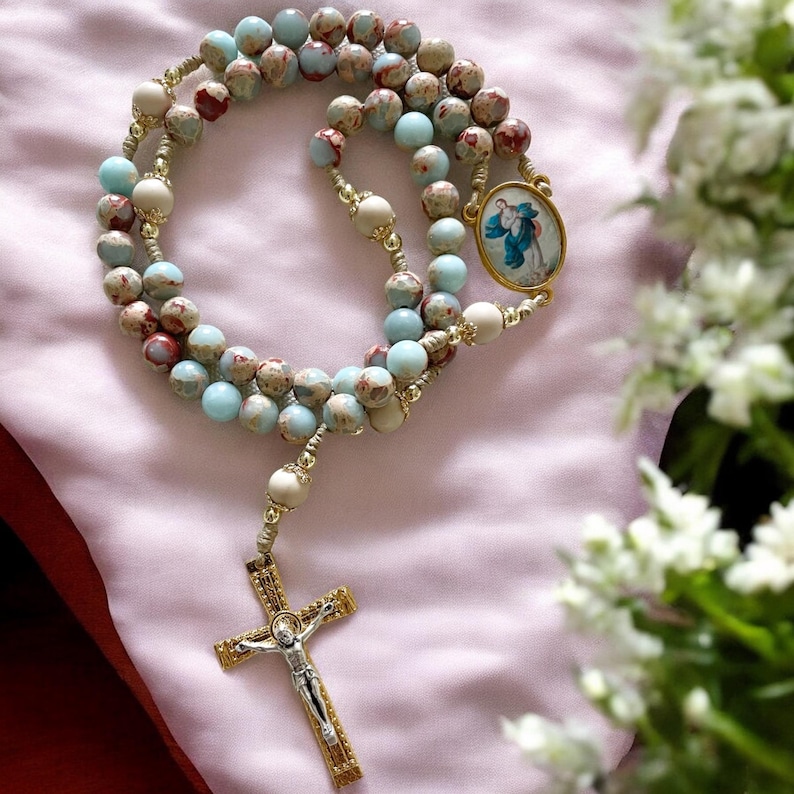 May include: A blue and brown rosary with a gold cross and a round pendant featuring a religious image. The rosary is on a pink fabric background.