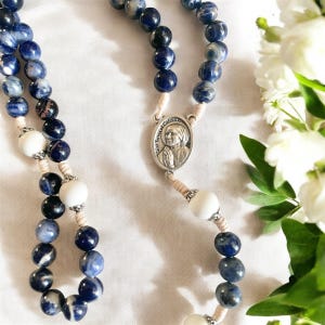 May include: A rosary with blue and white beads. The beads are strung on a beige cord, with a silver pendant featuring an image of a saint. White flowers are in the background.