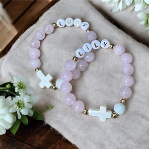 May include: Two pink and white beaded bracelets with gold accents. One bracelet has the name "LUCY" spelled out in white beads and the other has the name "LILY" spelled out in white beads. Each bracelet has a white cross charm.