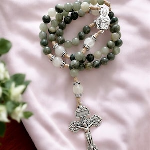 May include: A green and white stone rosary with a silver crucifix and a silver charm of Saint Anthony. The rosary is strung on a beige cord.