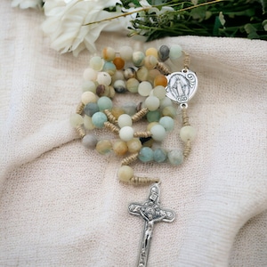 May include: A green, brown, and white beaded rosary with a silver cross and a silver charm featuring a religious image. The rosary is lying on a white fabric background.