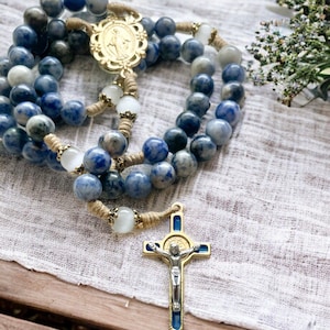 May include: A blue and white stone rosary with a gold-toned pendant and a silver-toned crucifix. The rosary is on a white fabric background.