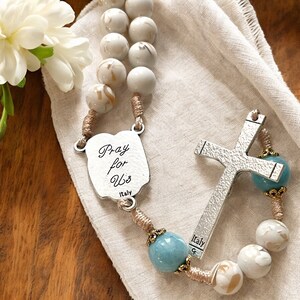 Puede incluir: Cuentas de rosario blancas y marrones con una cruz de plata y un dije de plata que dice "Pray for Us" con "Italy" debajo. Las cuentas est&aacute;n sobre un fondo de tela beige.