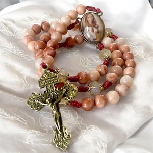 May include: A rosary with a red cord and a gold-toned crucifix. The beads are made of pink and white stone. The centerpiece features a round image of a woman in a white dress and a red scarf.