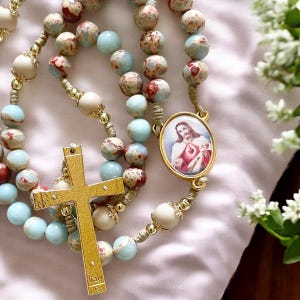 May include: A gold rosary with blue and red marbled beads, a gold cross, and a gold pendant with a picture of Jesus. The pendant has the words "Sacred Heart of Jesus" on it.