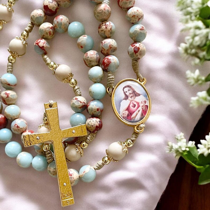 May include: A gold rosary with blue and red marbled beads, a gold cross, and a gold pendant with a picture of Jesus. The pendant has the words "Sacred Heart of Jesus" on it.