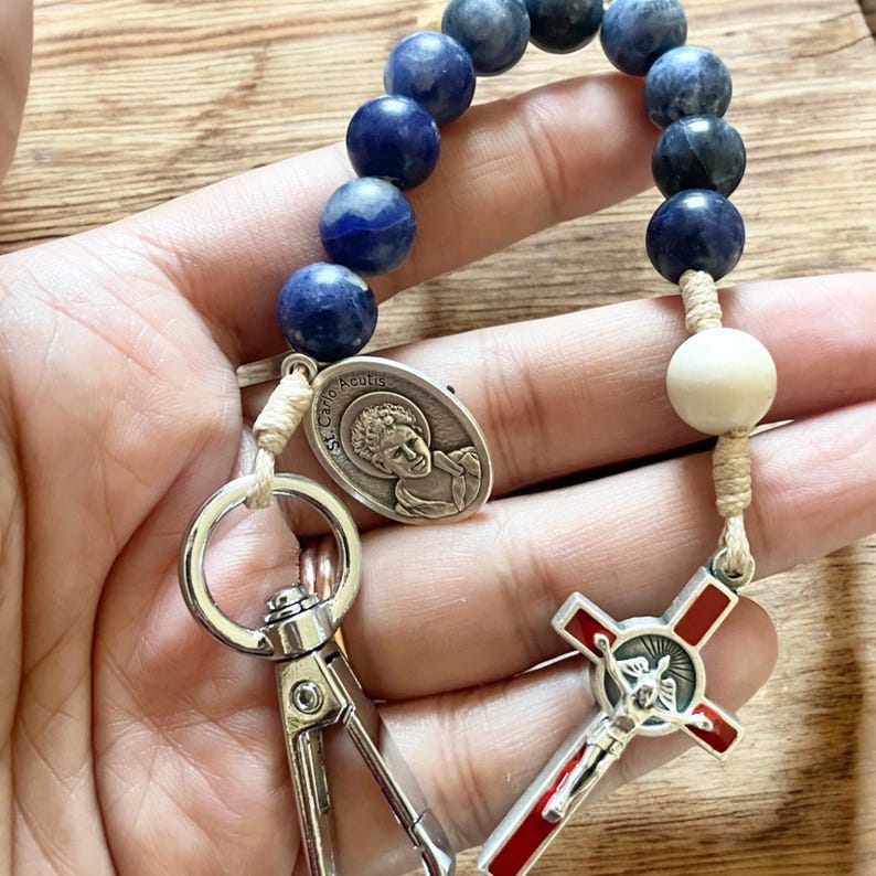 May include: A blue and white beaded rosary with a silver charm featuring a saint and a silver cross with a red enamel center. The rosary has a clasp for attaching to a keychain.