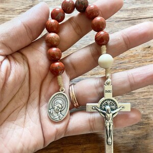 May include: A red stone rosary with a silver crucifix and a silver medal of Saint Carlo Acutis. The crucifix has the inscription "INRI" on the top.