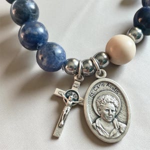 May include: A beaded bracelet featuring blue and cream-colored stone beads. A silver charm with a crucifix and a pendant with the text "St. Carlo Acutis" are attached to the bracelet.