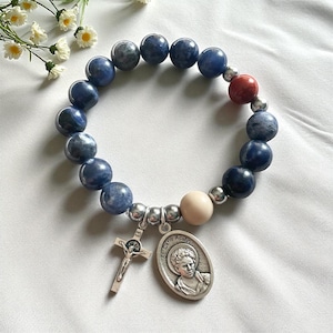 May include: A beaded bracelet featuring blue and gray round beads, a red bead, and a cream-colored bead. The bracelet includes a silver-toned crucifix charm and a St. Carlo Acutis pendant. The bracelet is displayed on a white surface.