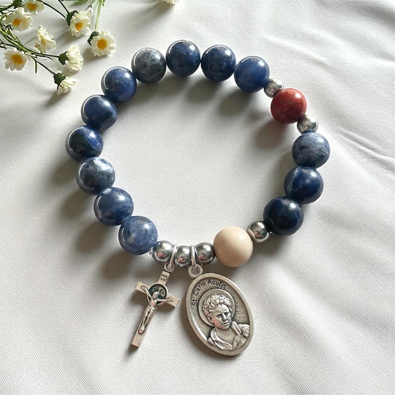 May include: A beaded bracelet featuring blue and gray round beads, a red bead, and a cream-colored bead. The bracelet includes a silver-toned crucifix charm and a St. Carlo Acutis pendant. The bracelet is displayed on a white surface.